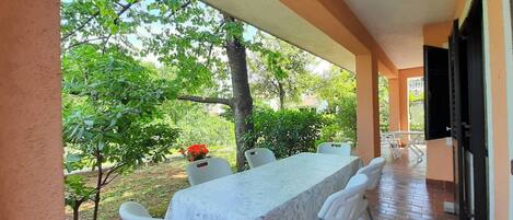 A covered patio with a table and chairs, a potted plant, and a view of greenery.