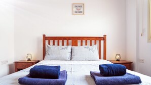 A neatly made bed with a wooden headboard, two pillows, and two folded towels. There are two bedside tables with lamps and a framed sign on the wall. - G402 Park View (Peyia Paphos)