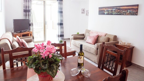 A living room with a dining table, chairs, a TV, and a cityscape painting on the wall. - G402 Park View (Peyia Paphos)