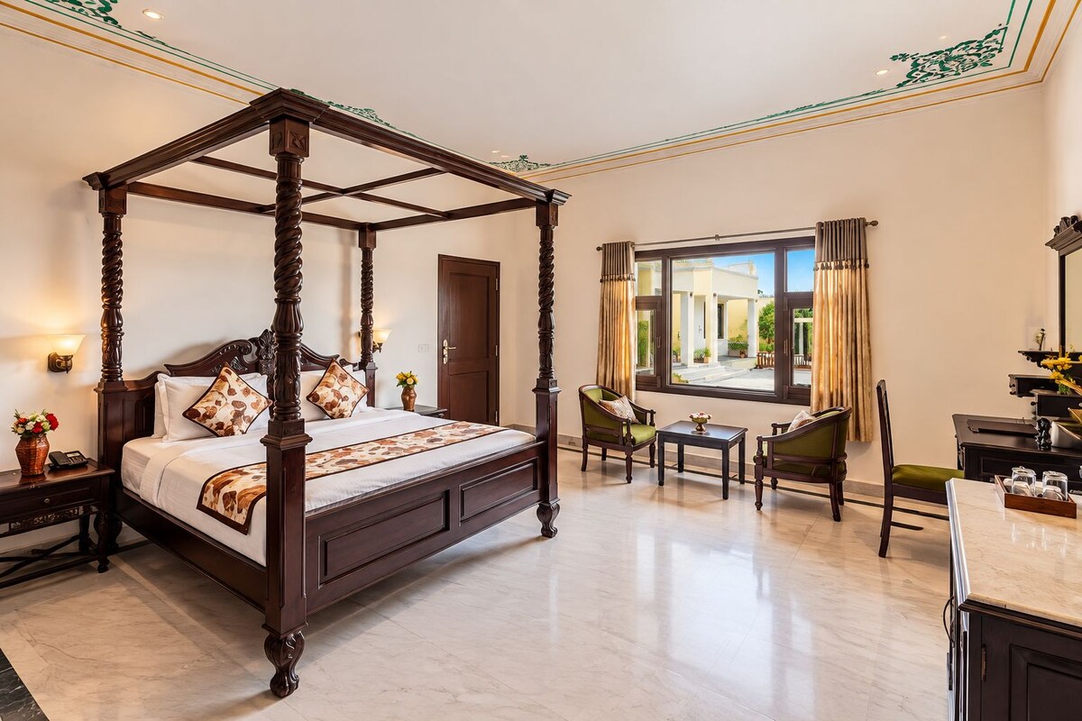 A four-poster bed with a marble floor, a sitting area with chairs and a table, and a view of the outside through a large window.