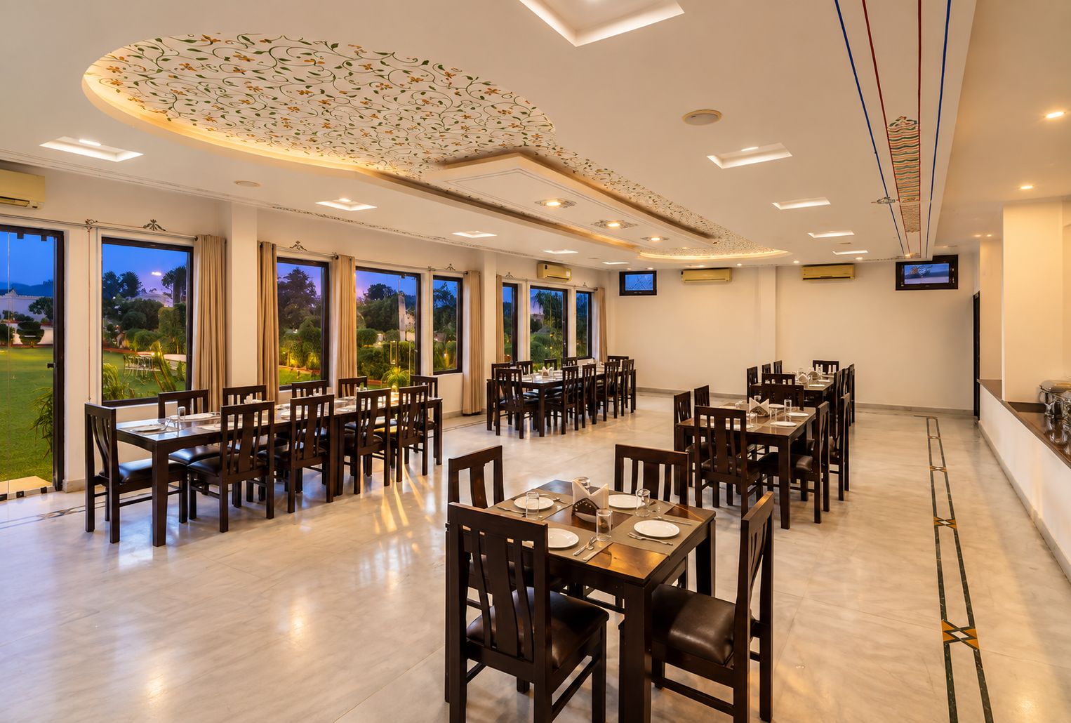 A spacious dining area with multiple tables and chairs, large windows offering a view of the outdoors, and a modern ceiling design.