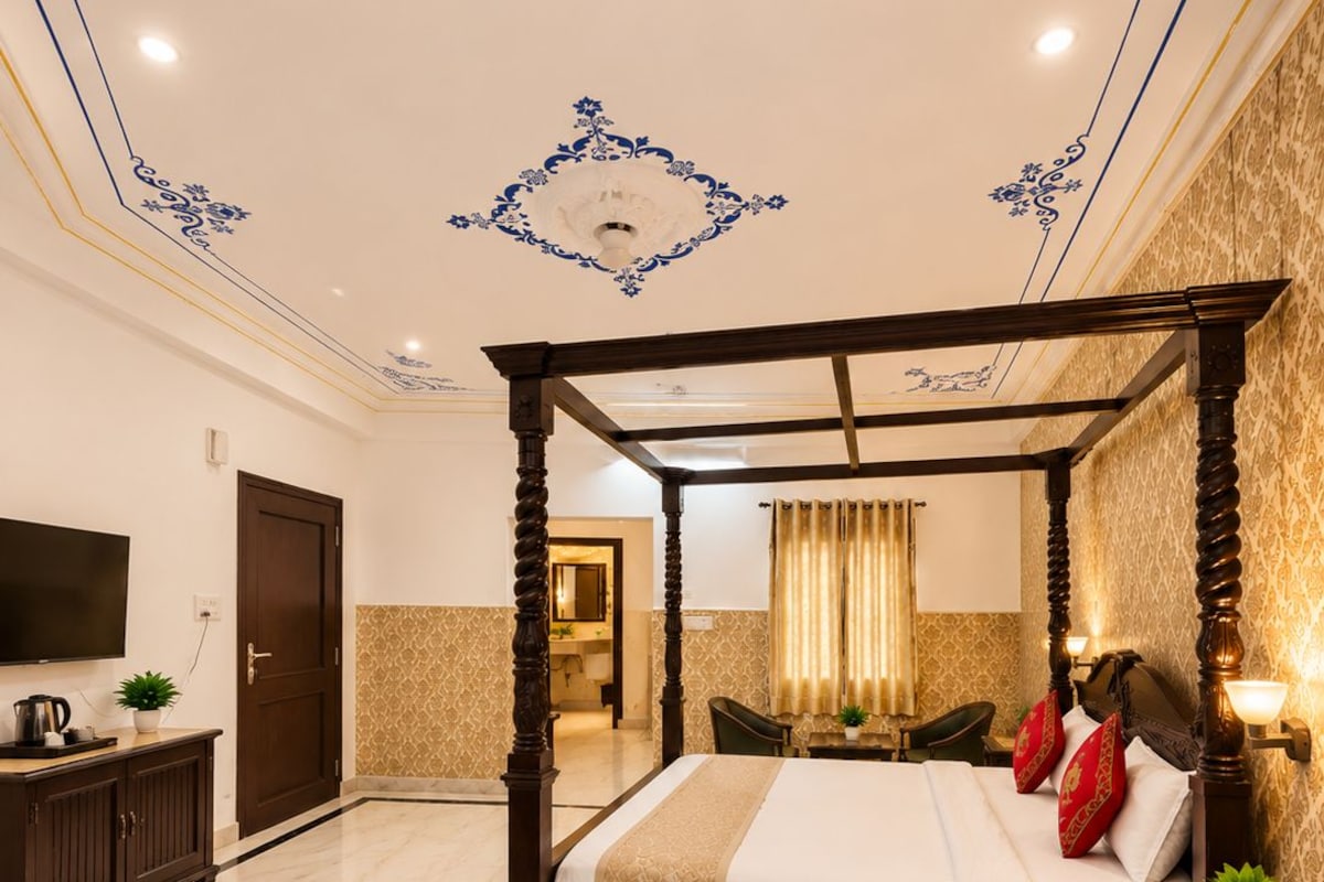 A four-poster bed with a headboard, a television on the wall, a desk with a chair, and a bench.