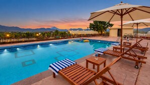 Zona de piscina con sillones y sombrillas, rodeada por una valla y vistas a las montañas.