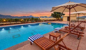 A pool area with lounge chairs and umbrellas, surrounded by a fence and a view of mountains. - Hill Garden Resort – Near Fateh Sagar, Udaipur (Udaipur)