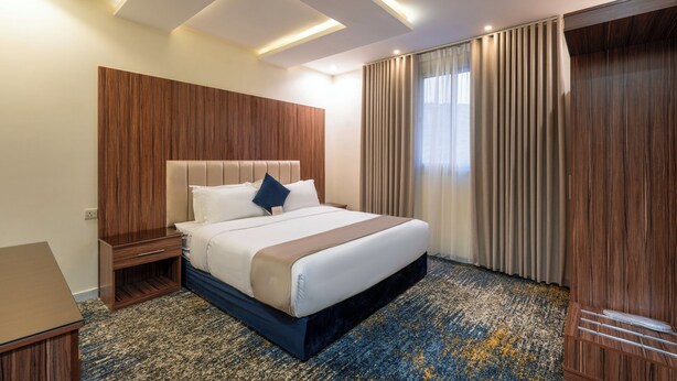 Habitación de hotel con cama, cabecero de madera, mesita de noche, escritorio y un gran ventanal con cortinas.