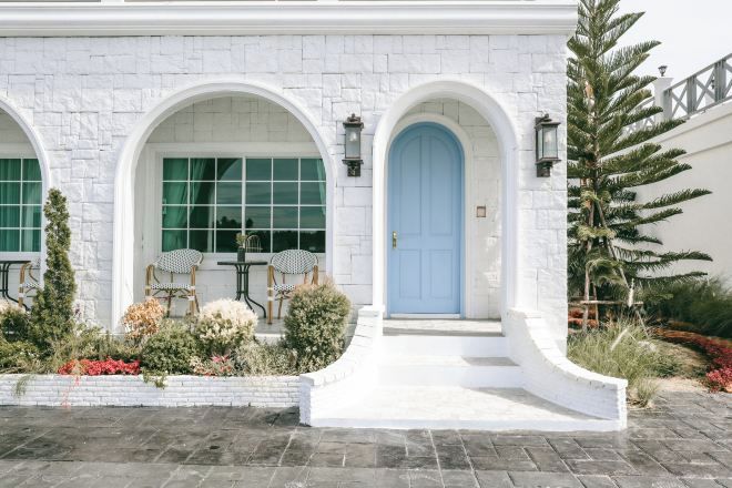 Un edificio blanco con ventanas arqueadas y una puerta azul, rodeado de un jardín ajardinado con diversas plantas y un pequeño espacio de descanso.