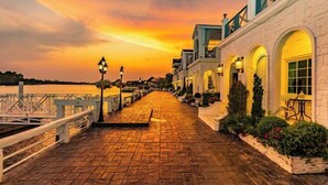 A waterfront promenade with a brick pathway, white buildings with arched windows, and street lamps. - Prima Amphawa (Mueang Samut Songkhram)