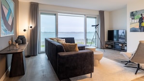 A modern living room with a sofa, a chair, a TV, and a large window overlooking a body of water. - Apartment in Zeeland by Oosterschelde Beach (Sint-Annaland)