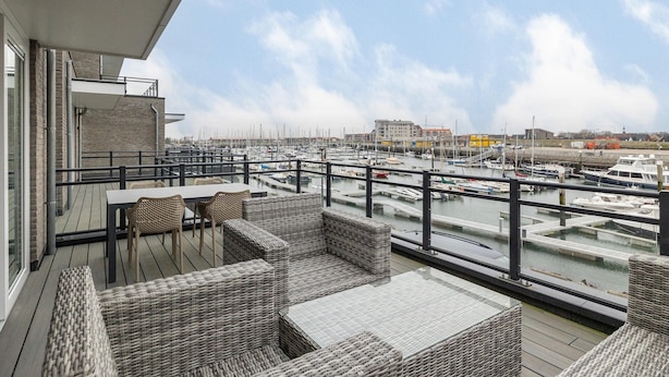 A balcony with wicker furniture overlooking a marina with boats. - Apartment in Zeeland by Oosterschelde Beach (Sint-Annaland)