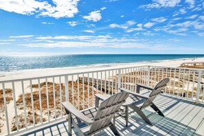 Una terraza de madera con dos sillas Adirondack con vista a una playa con valla blanca y dunas.