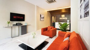 A modern lobby with a flat-screen TV displaying 'NETFLIX', a reception desk, and a seating area with orange sofas.