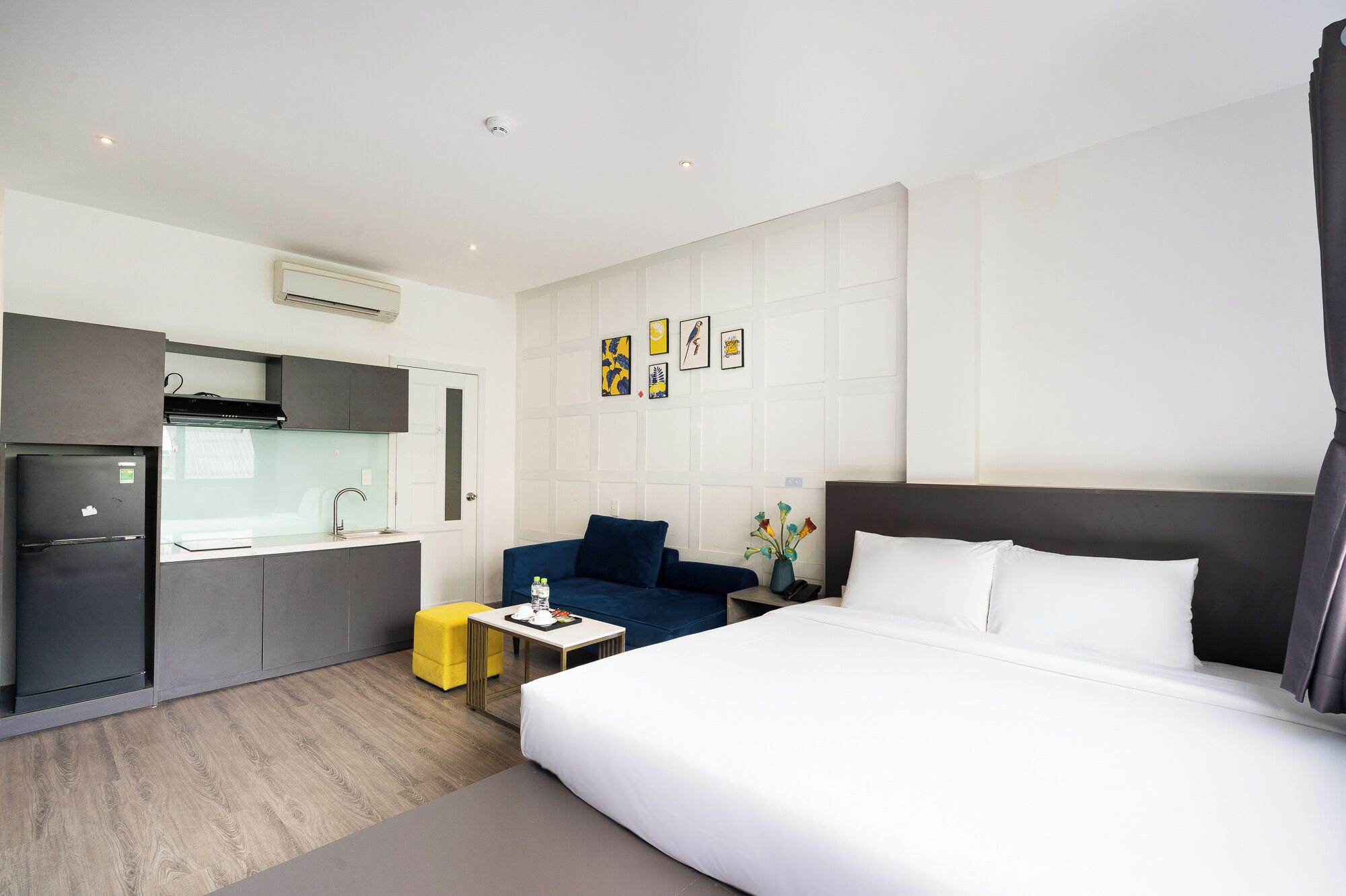 Una habitación de hotel moderna con una cama grande, una kitchenette pequeña, un sofá azul y una mesita de noche con un florero.