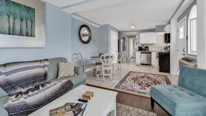 Ein modernes Wohnzimmer mit einer Couch, einem Couchtisch und einem Fernseher. Es gibt einen Essbereich mit Tisch und Stühlen. Im Hintergrund ist die Küche zu sehen.