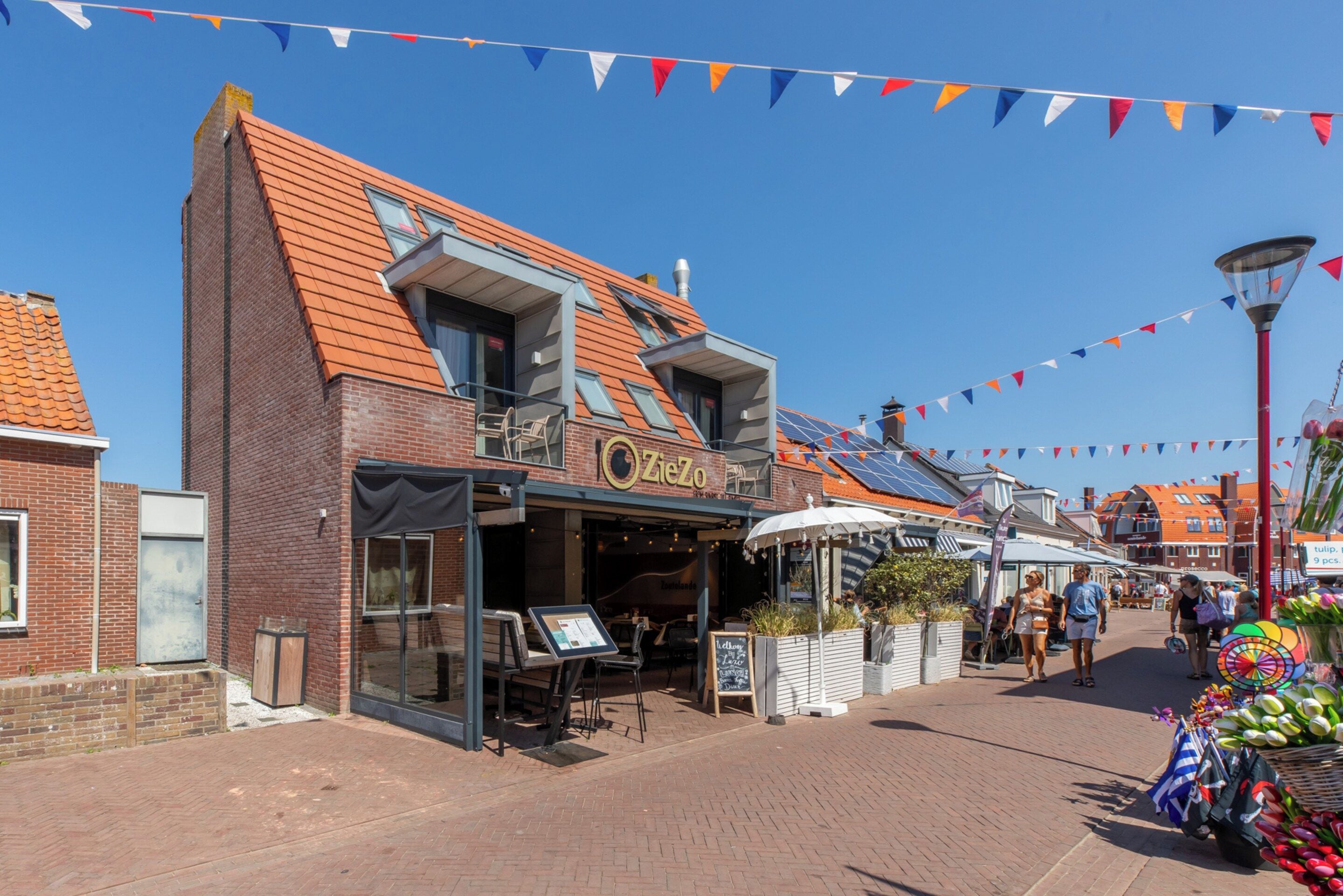 A brick building with a sloped roof, a storefront with outdoor seating, and a street with colorful flags.