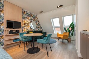 En moderne stue med et rundt bord, teal farvede stole, en grøn sofa, et fladskærms-tv og et stort spejl.