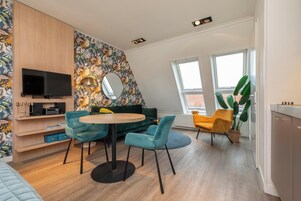 Ein modernes Wohnzimmer mit einem runden Tisch, blaugrünen Stühlen, einem grünen Sofa, einem Flachbildfernseher und einem großen Spiegel.