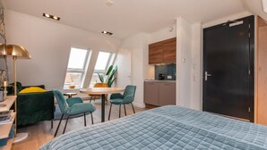 A compact bedroom with a large bed, a desk with a lamp, a chair, and a small dining table with chairs. There is a window providing natural light and a view of the outside. - Cozy Couple's Escape (Zoutelande)