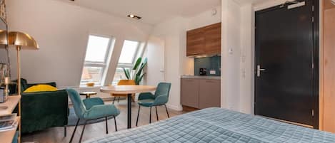 A compact bedroom with a large bed, a desk with a lamp, a chair, and a small dining table with chairs. There is a window providing natural light and a view of the outside.