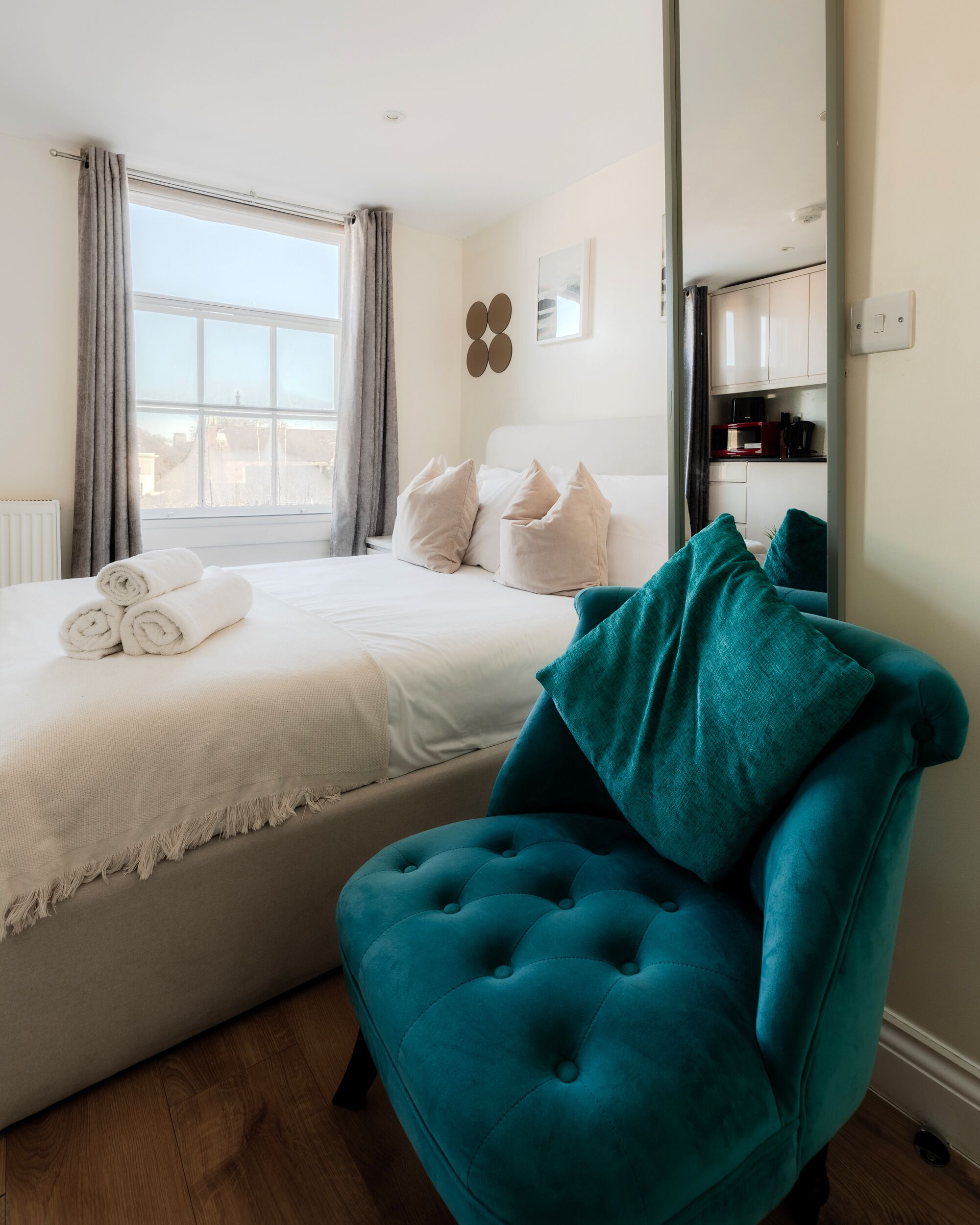 A bedroom with a bed, a teal armchair, a window with curtains, and a view of the city.