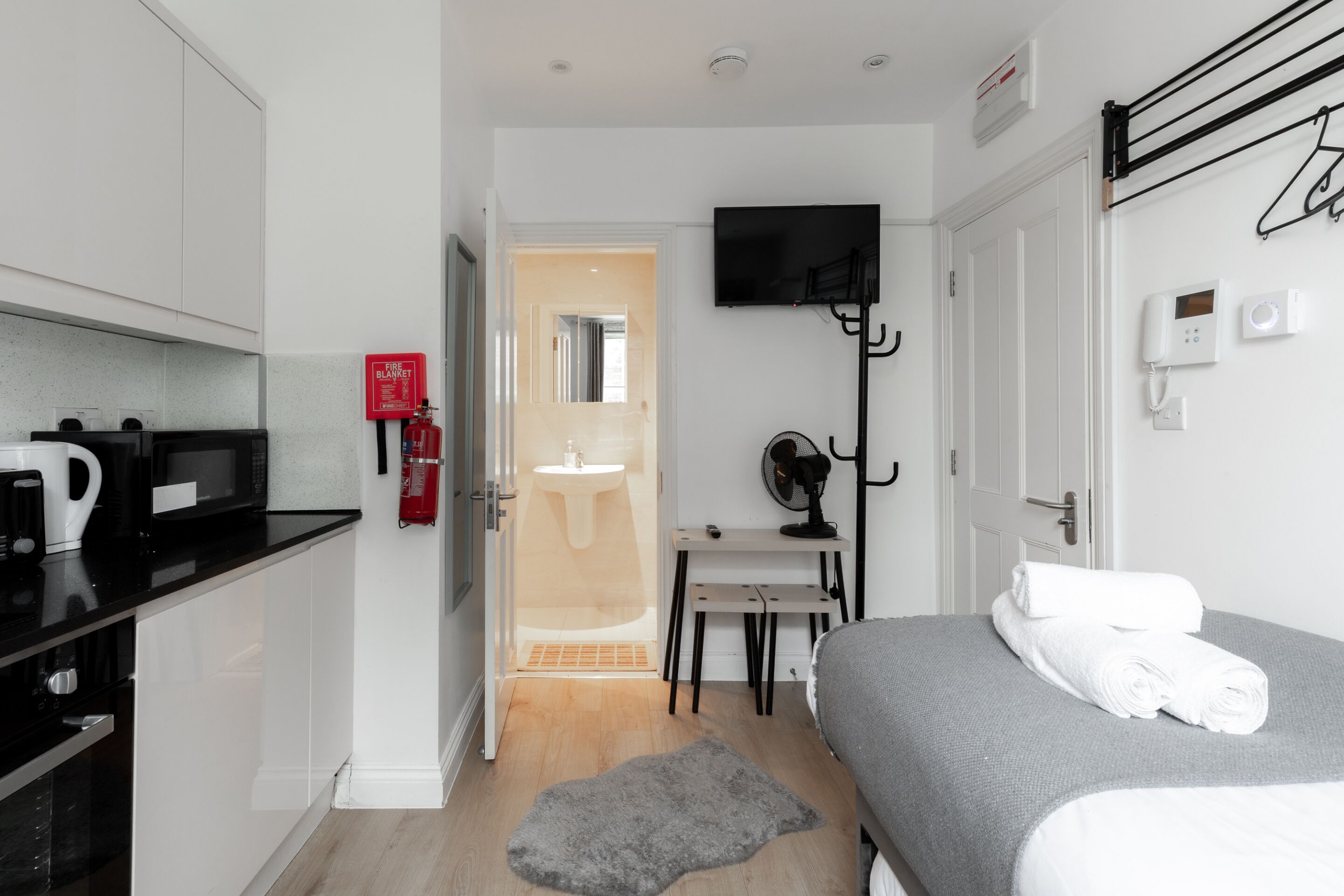 A modern hotel room with a kitchenette, a bed with neatly folded towels, a TV mounted on the wall, and a small table with a chair.