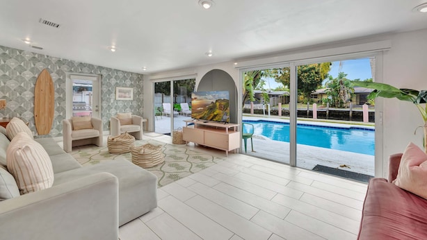 A modern living room with a pool view, featuring sofas, a TV, and a surfboard. - Poolside Paradise Dock & Pool! (Oakland Park)