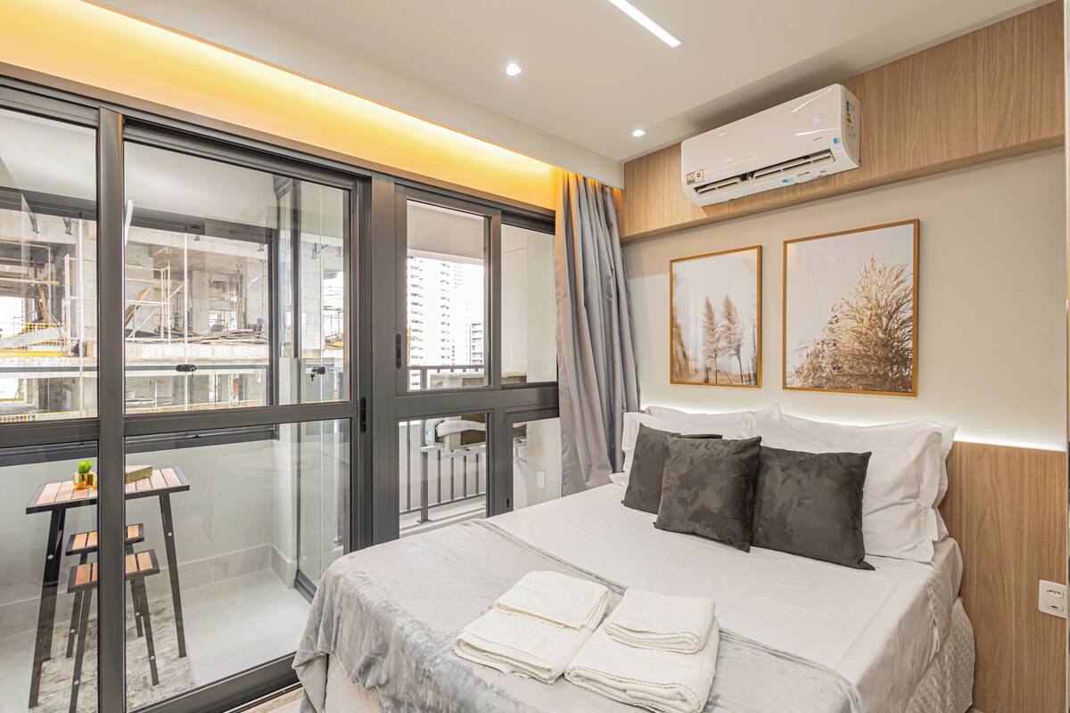 A modern hotel room with a large bed, a desk, and a balcony with a view of the city.