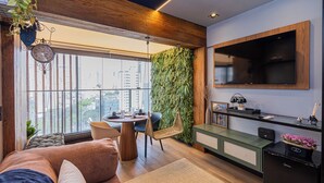 A modern living room with a large window, a dining area, a TV mounted on the wall, and a green vertical garden. - SSP - Soma Perdizes (São Paulo)