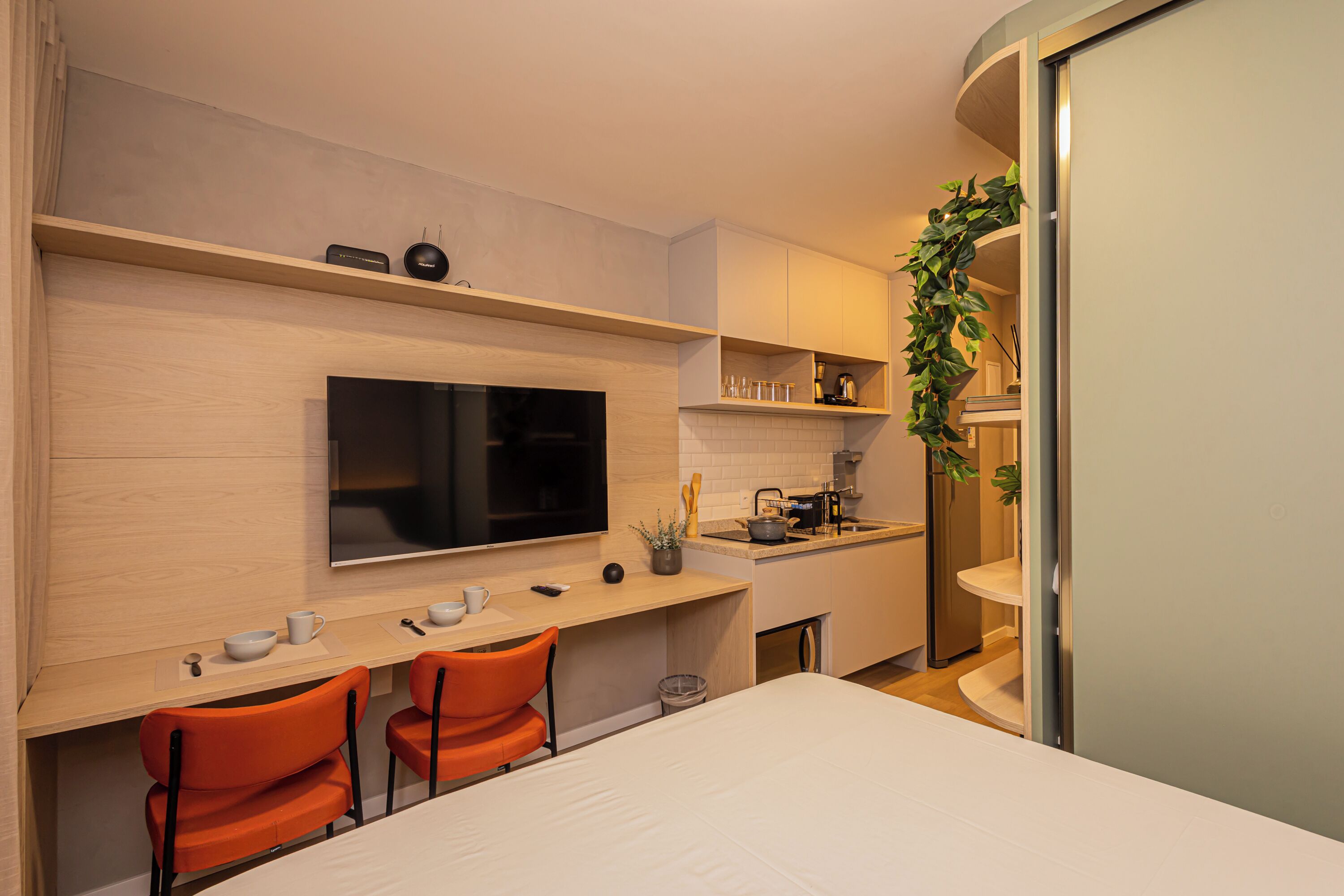 A modern kitchen with a flat-screen TV, a dining area with two orange chairs, and a small sink.