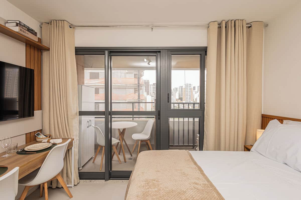 A hotel room with a bed, a desk with a chair, a small table with two chairs, a TV, and a balcony with a city view.