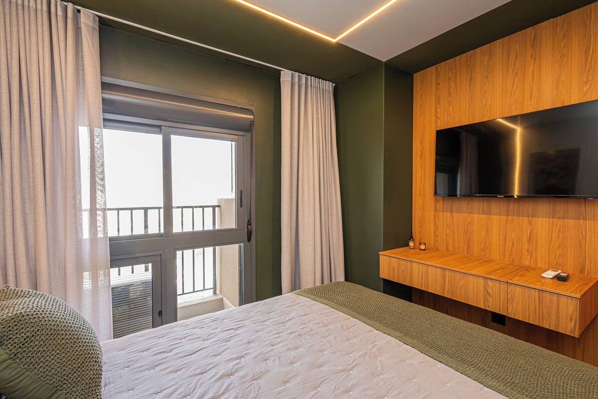 A hotel room with a bed, a TV mounted on a wooden panel, a window with curtains, and a balcony view.