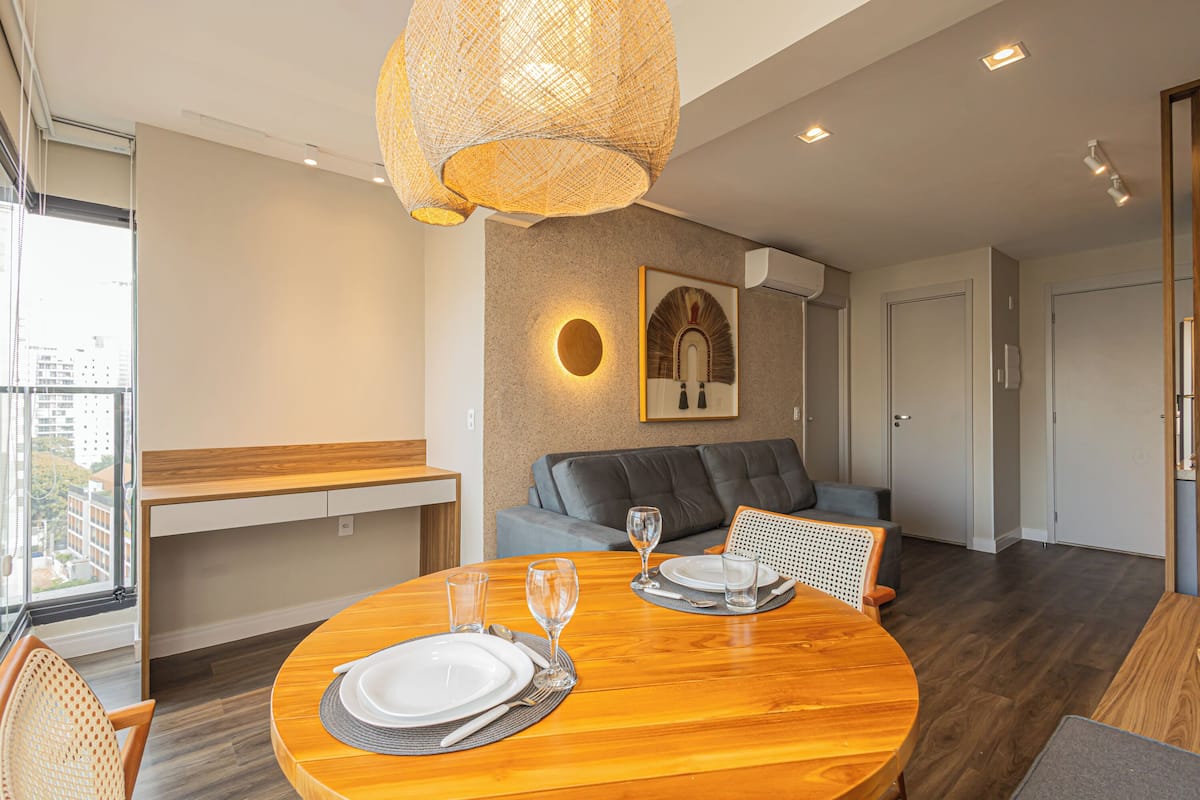 A modern dining area with a round wooden table set for two, a woven pendant light, a grey sofa, and a framed artwork on the wall.