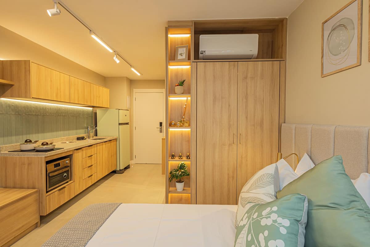 A modern bedroom with a bed, pillows, and a nightstand. Adjacent to the bedroom is a kitchen with wooden cabinets and a built-in oven.