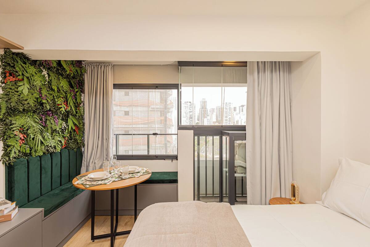 A hotel room with a bed, a small table with a breakfast set, a green cushioned bench, a balcony with a city view, and a green vertical garden.