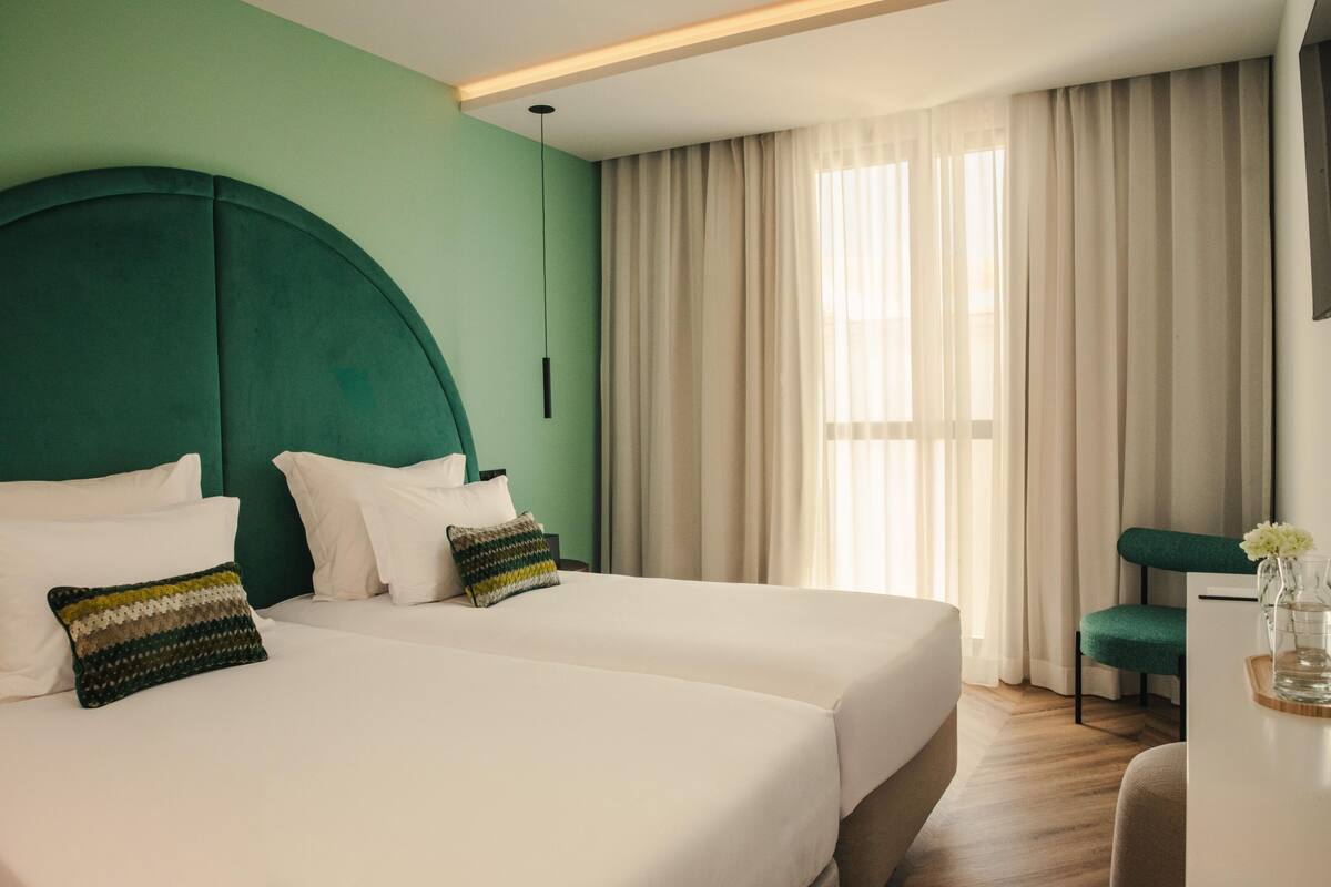 Une chambre d’hôtel avec deux lits, une tête de lit verte, du linge de lit blanc, un bureau agrémenté d’un vase de fleurs et une grande fenêtre avec des rideaux.
