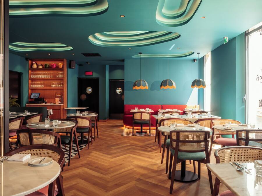 Ein Restaurant mit Holzböden, runden Tischen, Korbstühlen und einer blauen Wand mit Bücherregal.