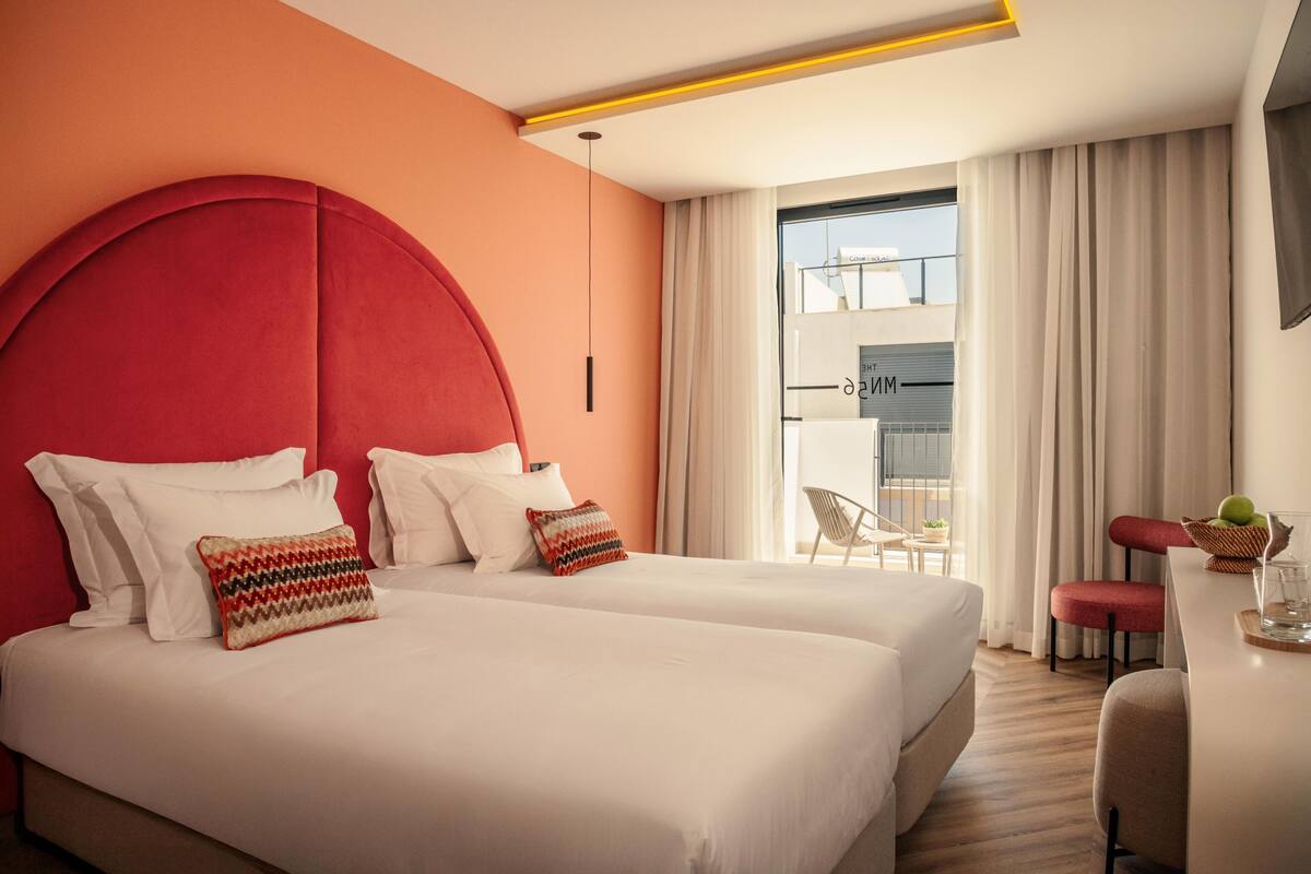 Une chambre d’hôtel avec deux lits, une tête de lit rouge, du linge de lit blanc, un coin repas avec une table et des chaises, et une vue sur l’extérieur.