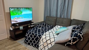 Un salón moderno con un televisor de pantalla plana sobre un mueble bajo, un sofá marrón, ropa de cama estampada y suelo de madera.