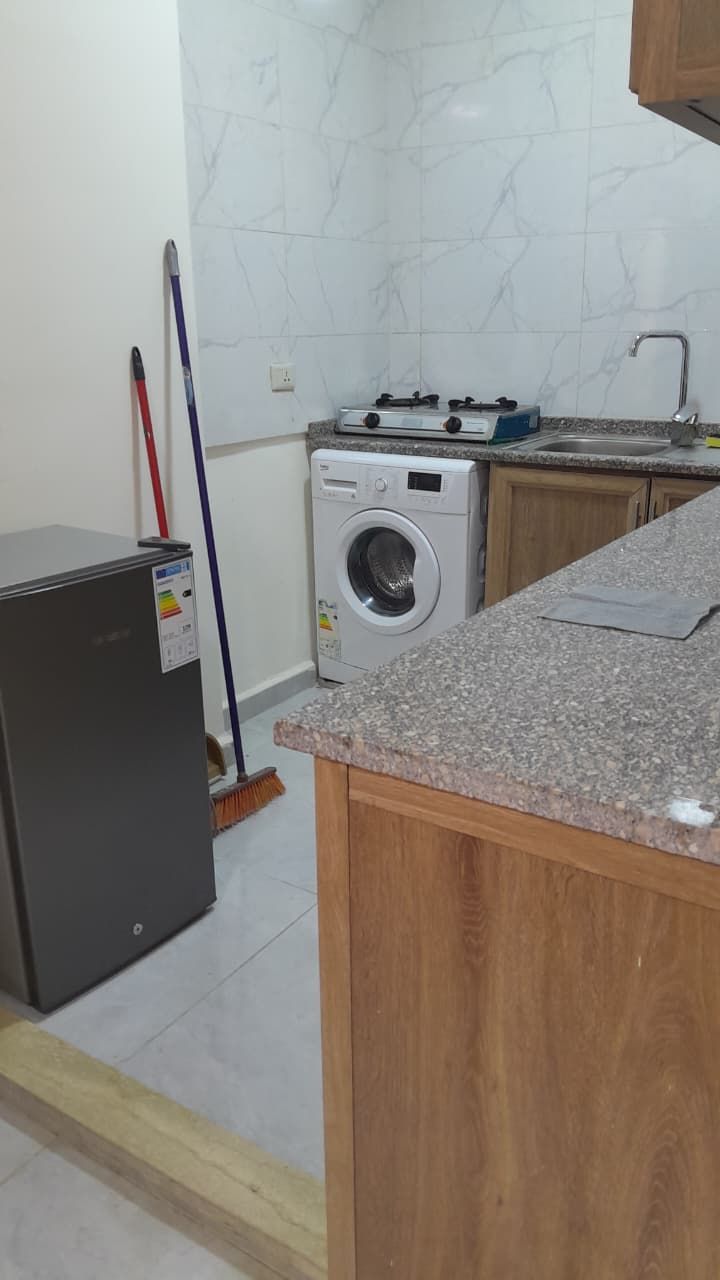 A kitchen with a washing machine, a small refrigerator, and a broom.