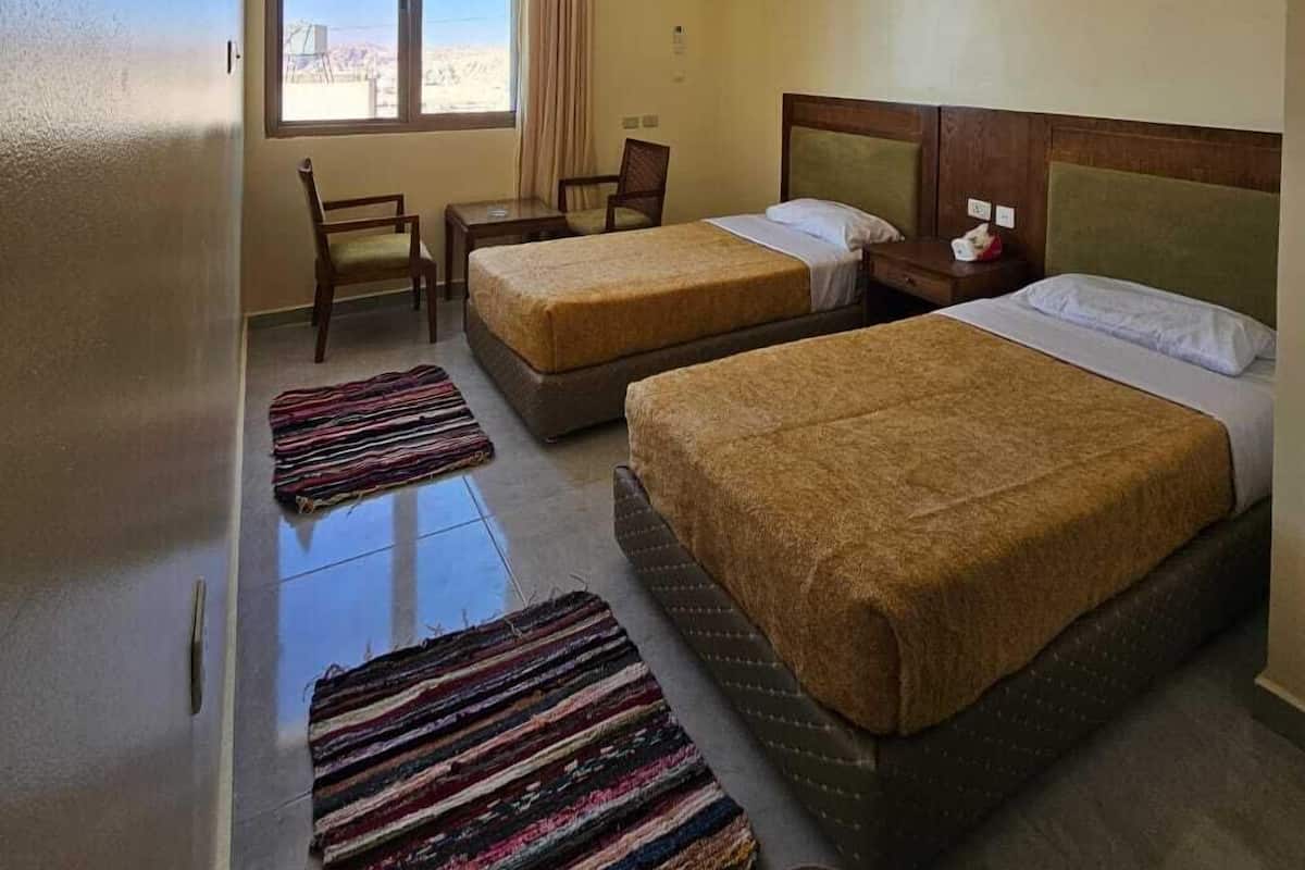 A hotel room with two beds, a window with curtains, a small table, a chair, and a colorful rug.