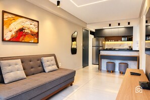 A modern living room with a grey sofa, a large abstract painting, and a kitchen area with dark cabinets and a stainless steel refrigerator. - Mardisa Design - LikeHome Hospedagens (João Pessoa)