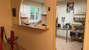 Une cuisine avec des meubles blancs, une fenêtre, un plan de travail sur lequel se trouvent divers objets, et un coin repas avec des chaises rouges.