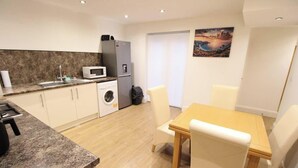 A compact kitchen with a washing machine, a microwave, and a refrigerator. There is a dining table with chairs and a painting on the wall.