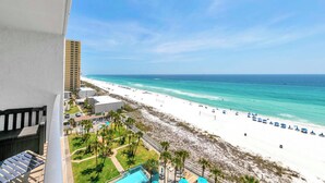 A beach view from a balcony overlooking a sandy beach with a pool and palm trees.