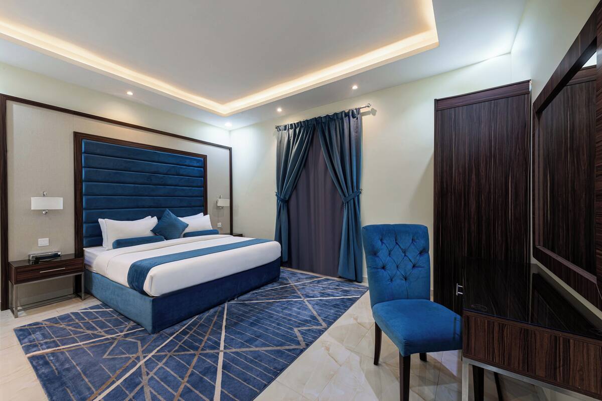 Une chambre d’hôtel moderne avec un grand lit, un fauteuil bleu rembourré, une armoire et un tapis à motifs.