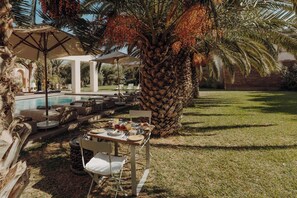 An outdoor dining area with a table set for two, surrounded by lounge chairs and a palm tree. - The Garden of Siredrah, an Oasis with Pool and Hammam (Marrakech)