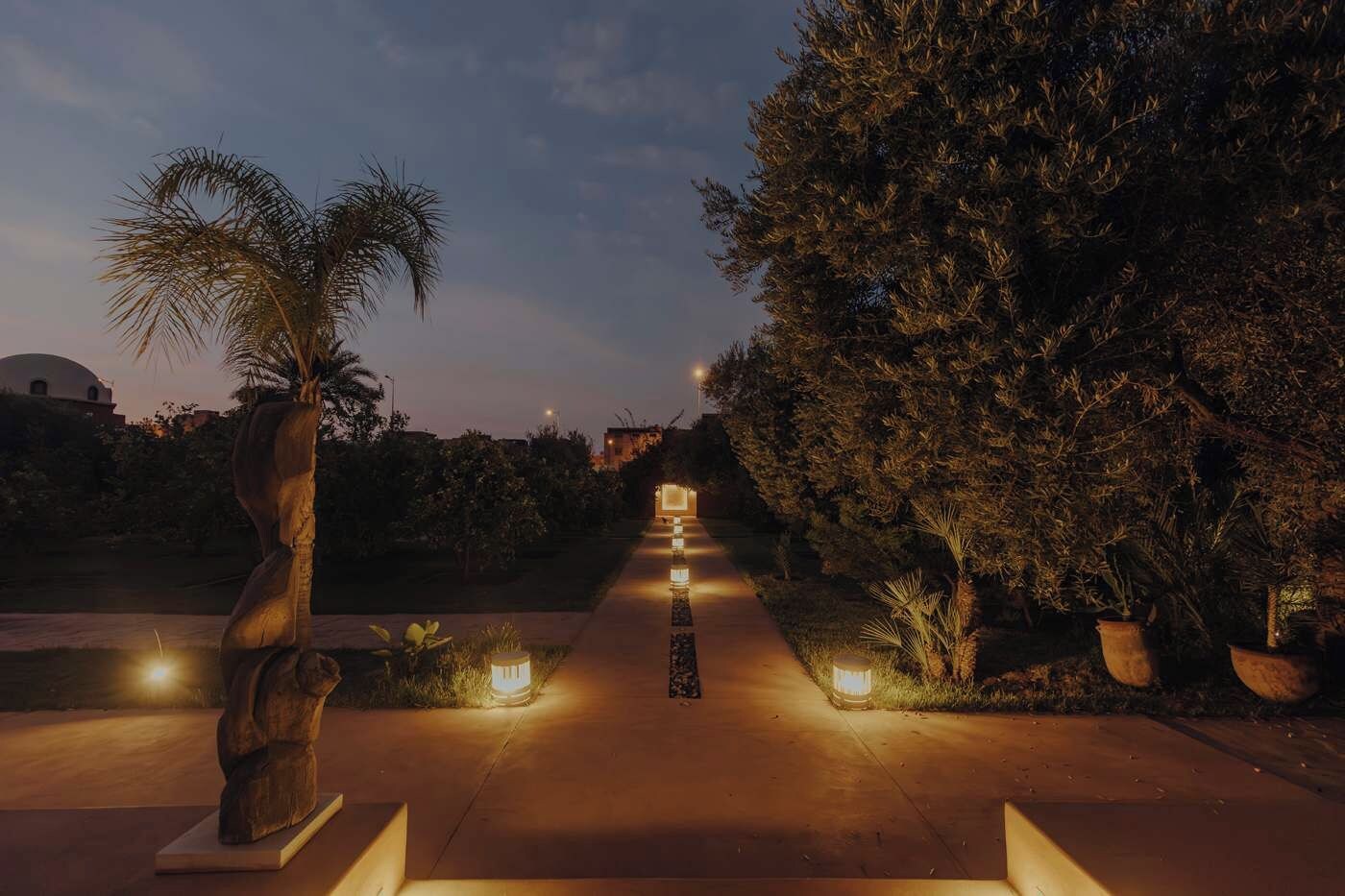Uma trilha bordeada por árvores e iluminada por luzes baixas, que conduz a um edifício com cúpula.