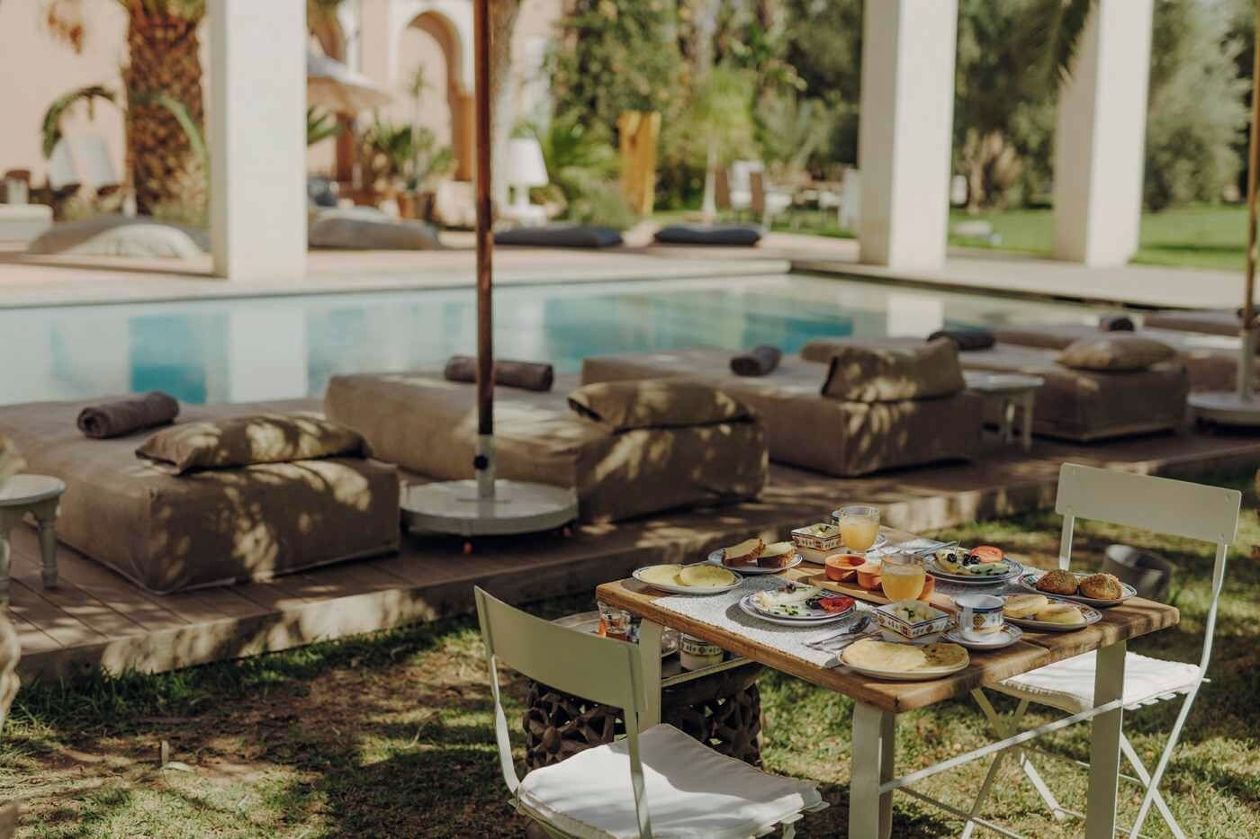 An outdoor dining area with a pool, cushioned seating, and a table set with food and drinks.