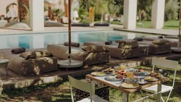An outdoor dining area with a pool, cushioned seating, and a table set with food and drinks.