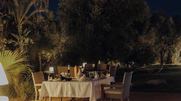 An outdoor dining area with a table set for two, surrounded by chairs, with a view of trees and a night sky.