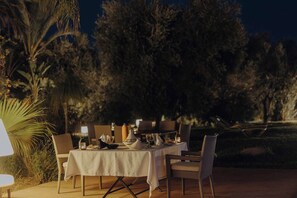 An outdoor dining area with a table set for two, surrounded by chairs, with a view of trees and a night sky.